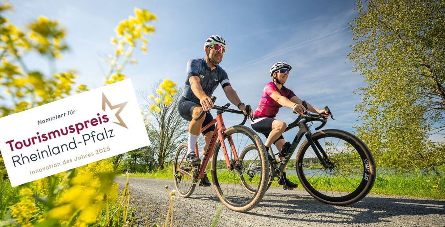 Zwei Radfahrer fahren auf einer sonnigen Landstraße, umgeben von grünen Feldern und gelben Blumen. Ein Schild in deutscher Sprache weist auf eine Nominierung für den rheinland-pfälzischen Tourismuspreis 2025 für die Innovation des Jahres hin.